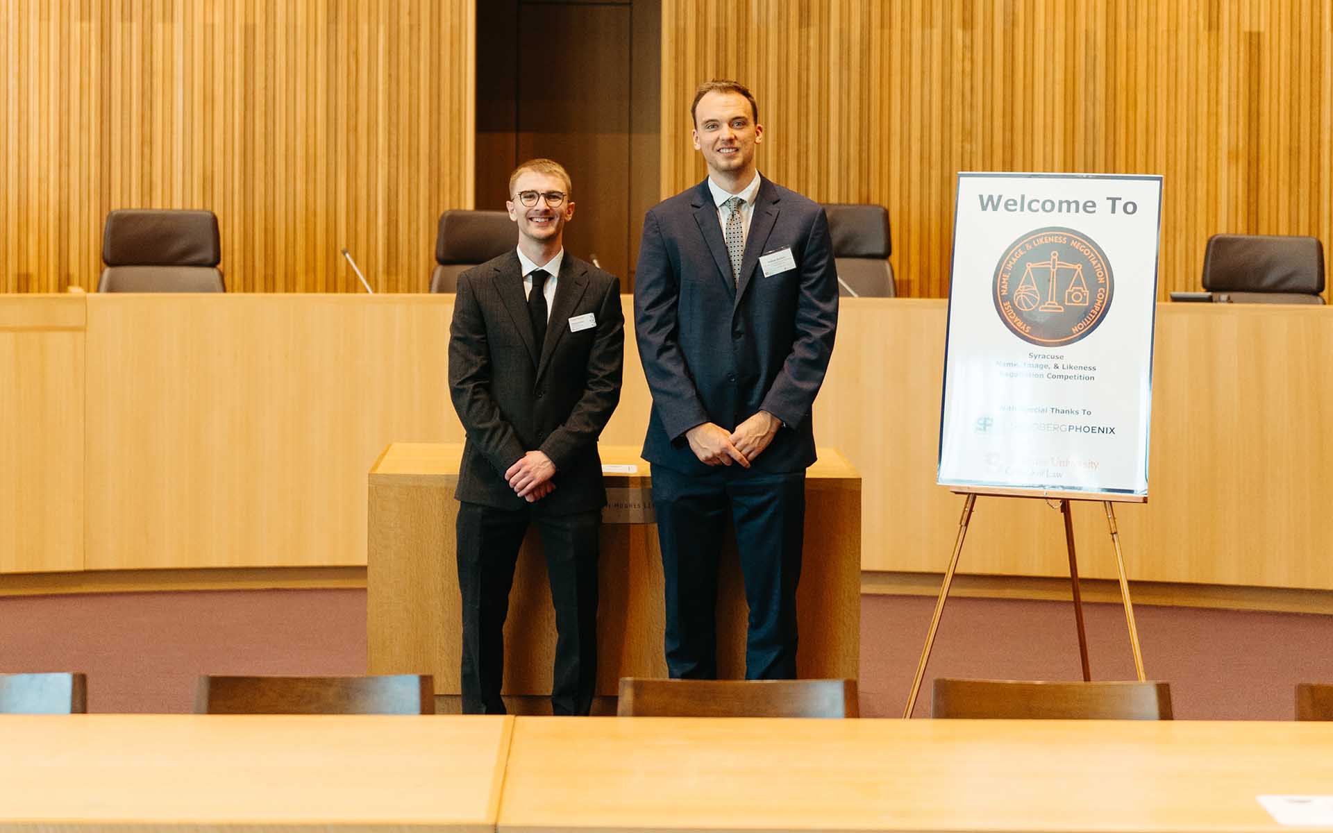 Two students stand in front of the Melanie Grey Courtroom next to a Welcome to the NIL Competition sign.