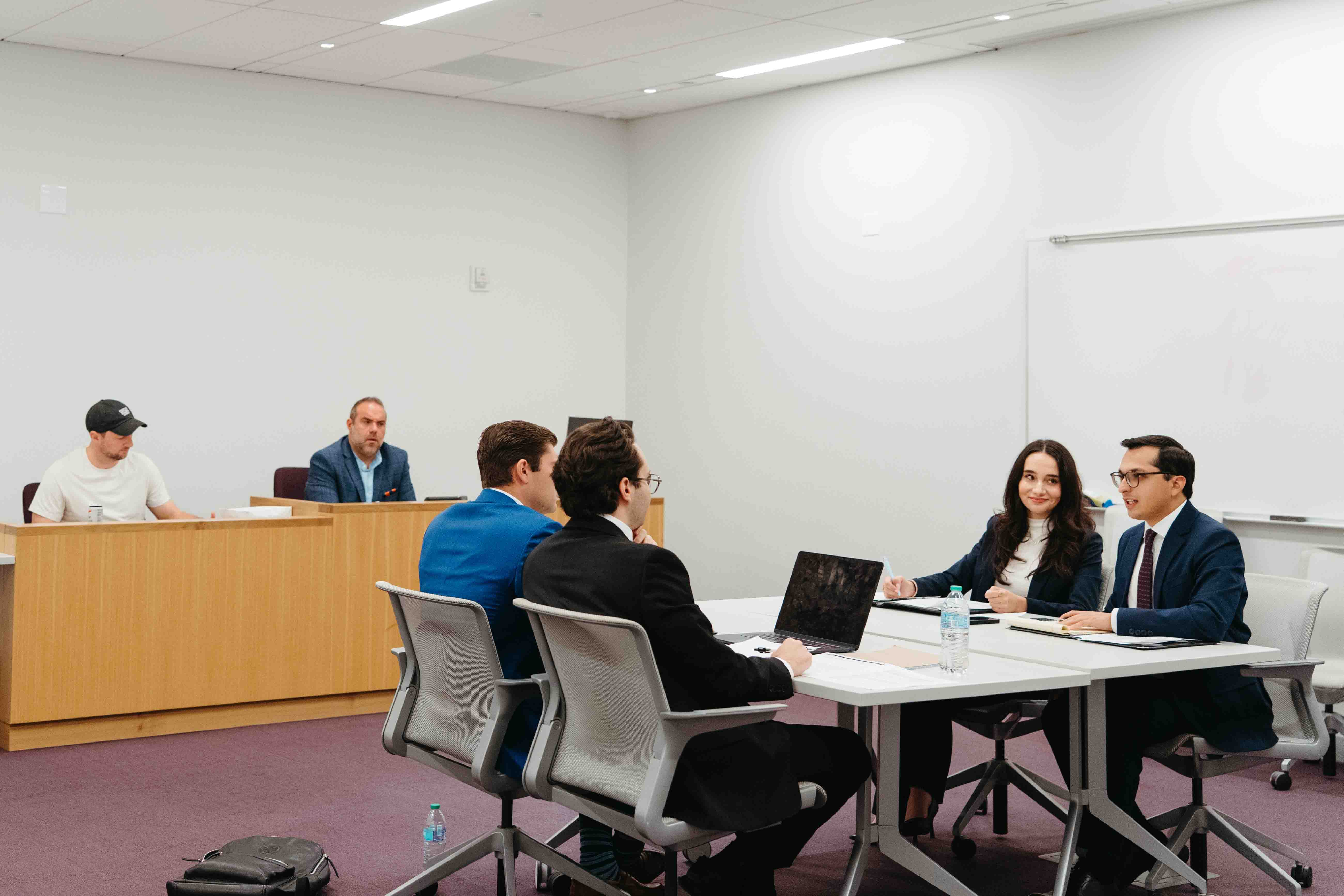 four students compete in an advocacy competition at the table in a room in front of two judges.