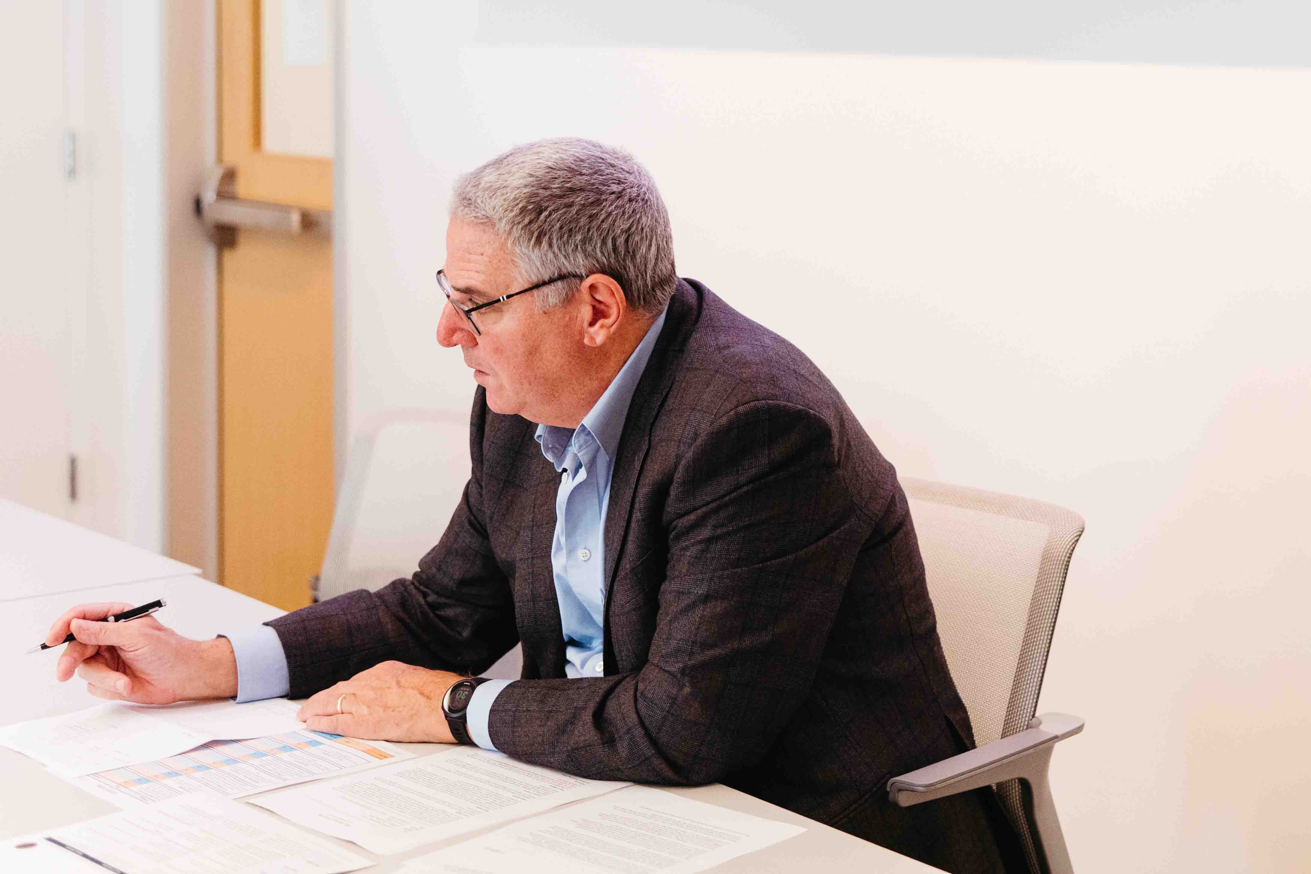 man sits at desk with a pen in front of papers, looking up and wearing glasses.