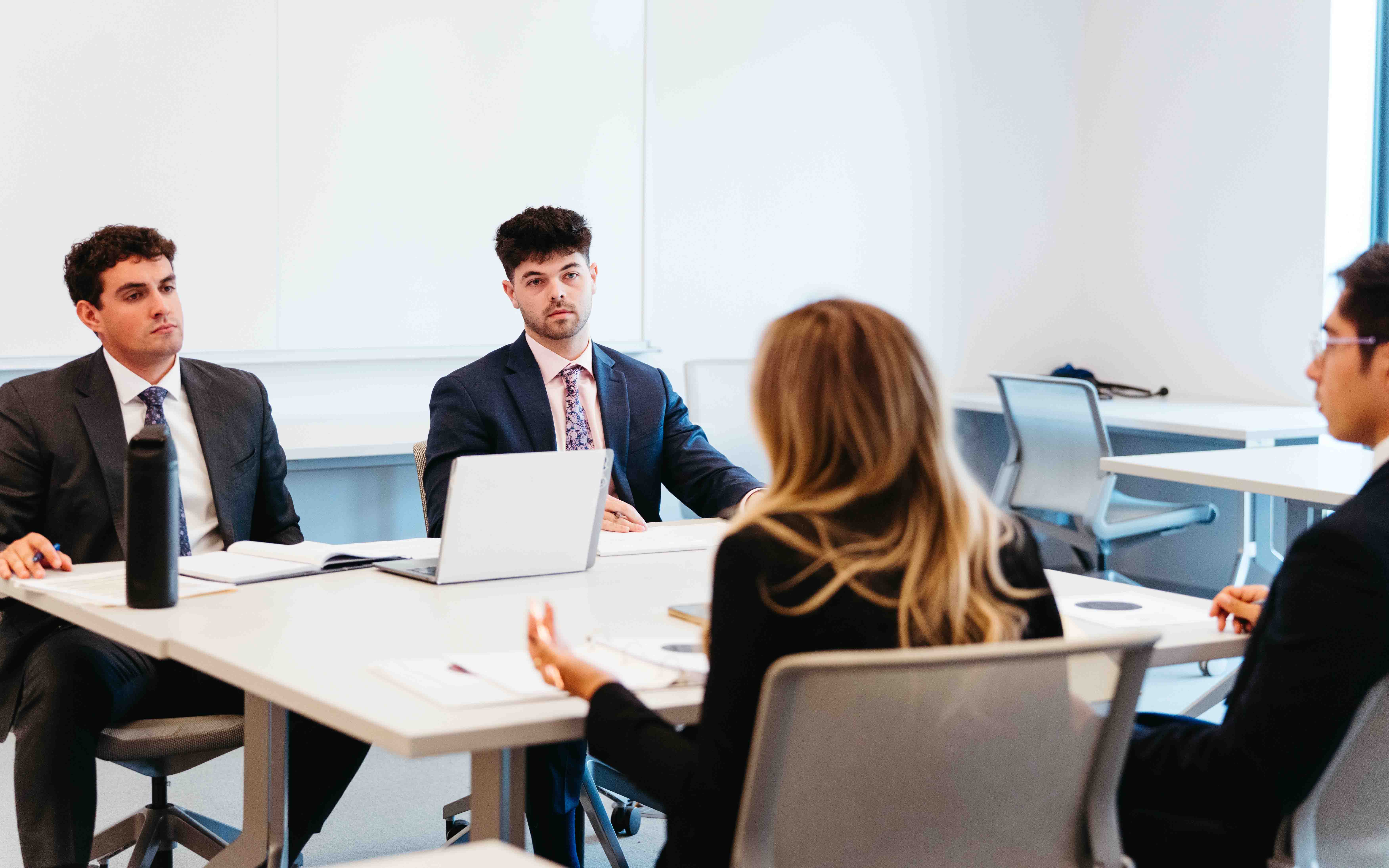 two students compete with two others sitting around a table at the NIL competition, wearing suits.