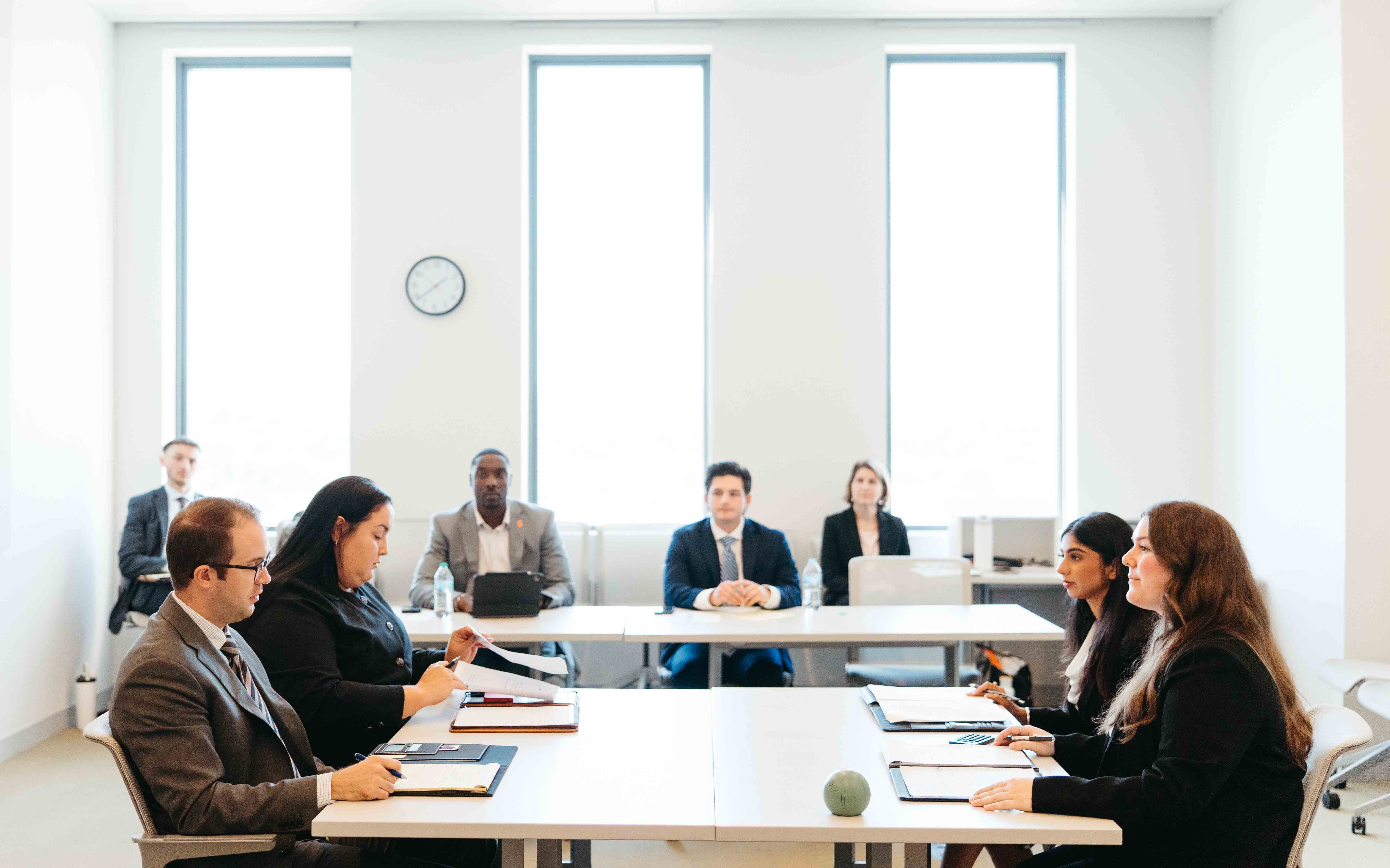Two competing teams at the NIL competition sit at a table with a room with three large windows in front of a judges panel.