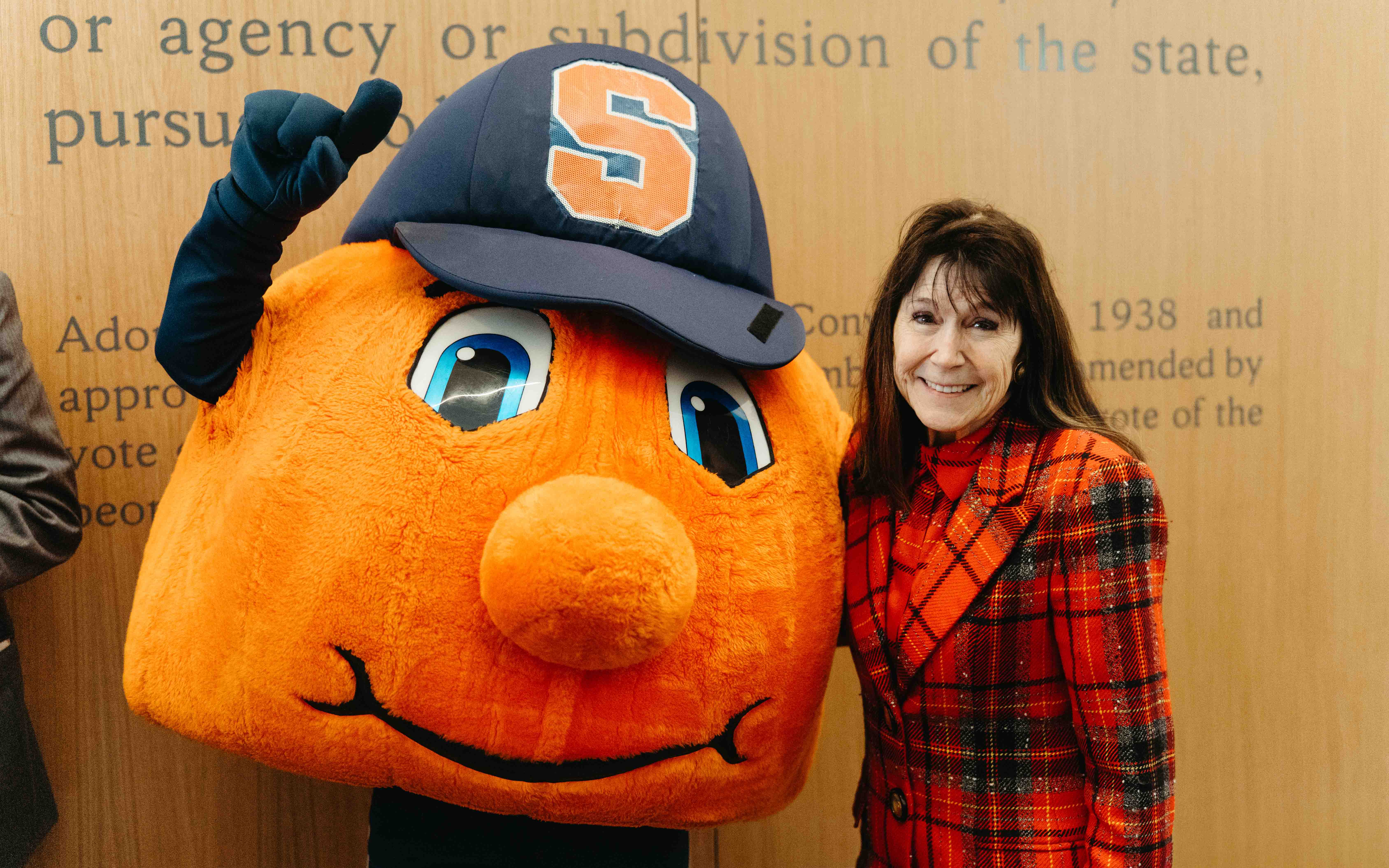 Jeanne Olivier smiles with Otto the Orange mascot at SU in front of a wooden wall.