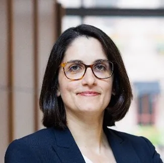 Headshot of Alia in front of abstract background, wearing brown rimmed glasses and a blue suit.