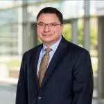 Headshot of man in dark suit and tie with dark hair and eyeglasses