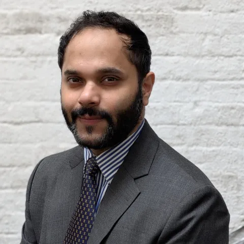 Headshot of Guarav in front of a white brick wall, wearing a gray suit.