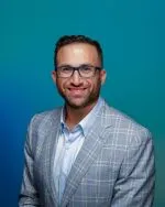 Headshot of man in grey checked suit with white shirt and short dark hair and beard wearing eyeglasses