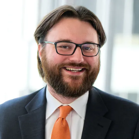 A man with dark hair and glasses wearing a dark suit and orange tie.