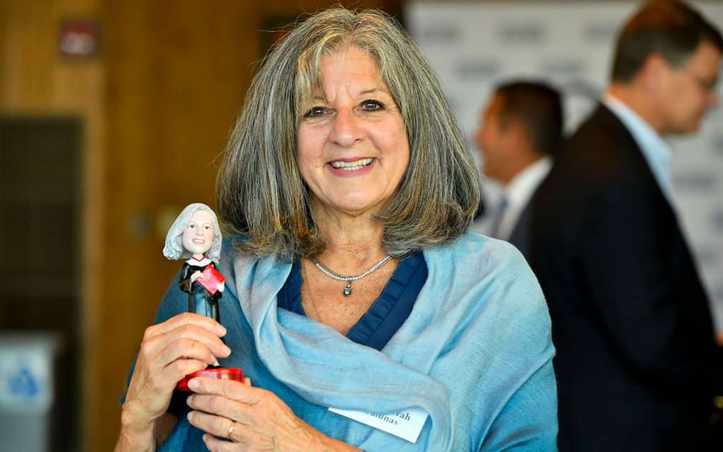 Judge Karalunas holds up a bobblehead of herself and smiles at the camera wearing a blue blouse