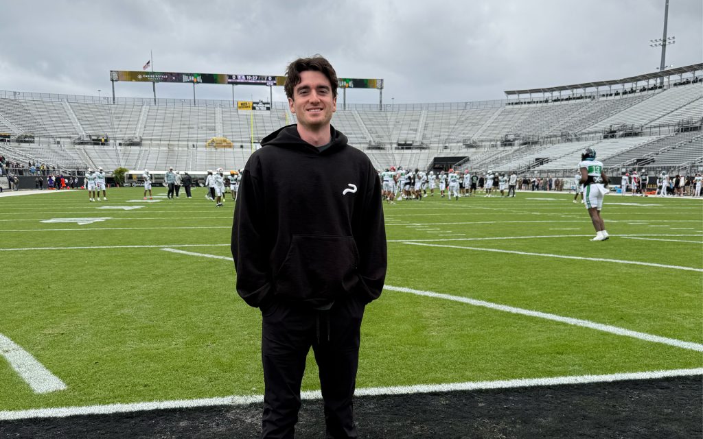 A young man with dark hair, wearing a black hoodie with a small white logo on the left chest, stands on a football field with a bright green turf. Behind him, football players in white and green uniforms are practicing, and a large stadium with empty stands is visible under an overcast sky.
