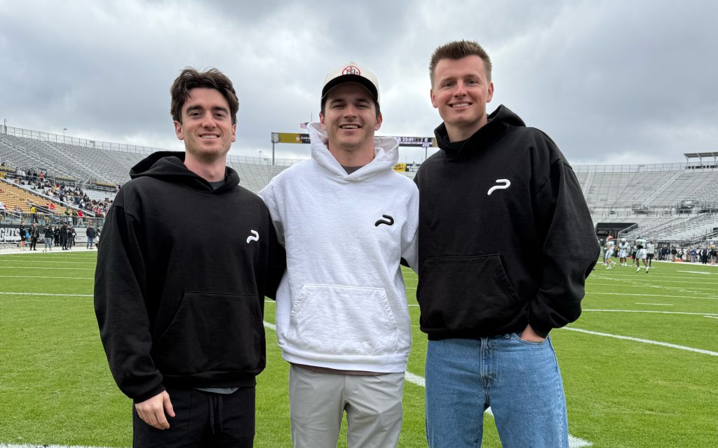 Three young men stand together on a football field, smiling at the camera. The two on the left and right wear matching black hoodies with a white logo, while the one in the middle wears a white hoodie and a baseball cap. Behind them, players are practicing on the field, and stadium stands with some spectators are visible under a cloudy sky.