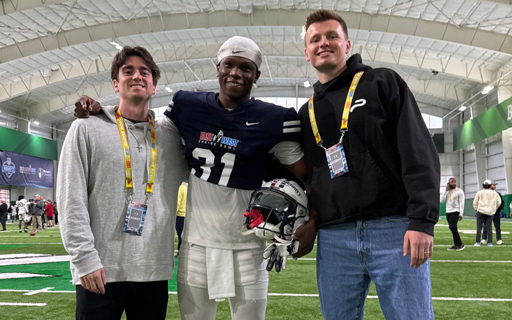 Two young men pose inside an indoor football training facility with bright green turf. One wears a light gray hoodie and has a credential badge around his neck, while the other wears a black hoodie with a white logo and also has a badge. Between them, a football player in a navy blue East-West Shrine Bowl uniform (#31) holds his helmet and smiles at the camera