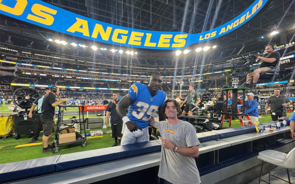 A young man in a gray Los Angeles Chargers t-shirt stands on the sidelines of an NFL stadium, pointing towards a football player in a blue and yellow Chargers uniform (#39). The player leans over the barrier, smiling. The stadium is brightly lit, with a large "Los Angeles" sign visible in the background and several media crew members working nearby.