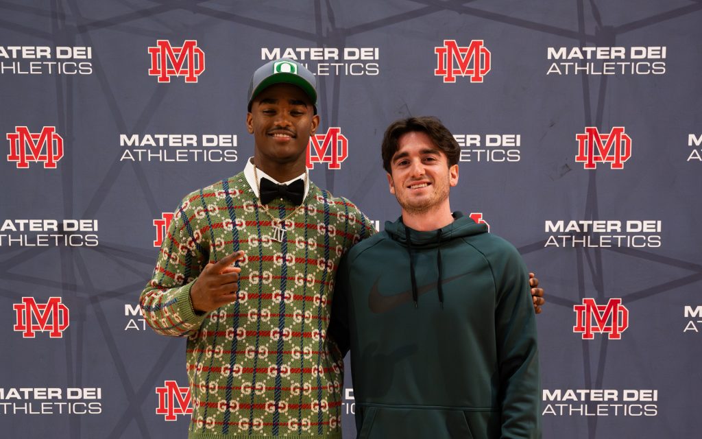 Two young men stand in front of a Mater Dei Athletics backdrop. The man on the left wears a stylish green Gucci sweater, a black bow tie, a gray Oregon Ducks cap, and a chain necklace with "NT" initials. He makes a hand gesture while smiling at the camera. The man on the right wears a dark green Nike hoodie and smiles.