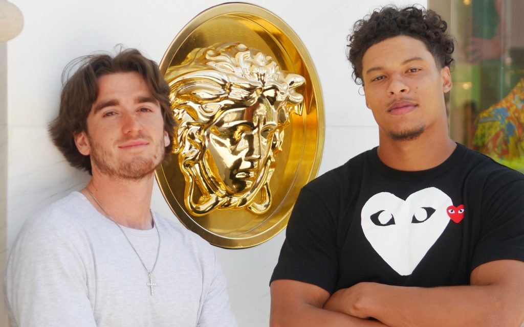 Two young men pose in front of a large golden Versace Medusa emblem. The man on the left has shoulder-length wavy brown hair, wears a light gray t-shirt and a silver cross necklace, and leans slightly towards the other. The man on the right wears a black t-shirt with a white heart-eyed logo and crosses his arms, looking directly at the camera.