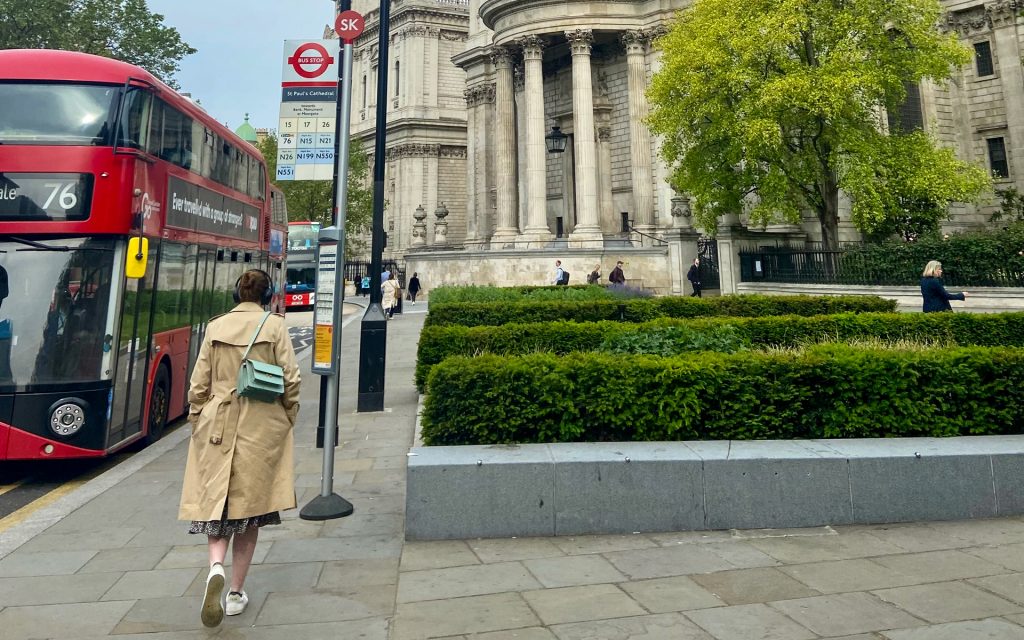 A person in a trenchcoat walks passed a double decker bus