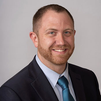 headshot of andrew lloyd smiling in a suit in front of a gray background