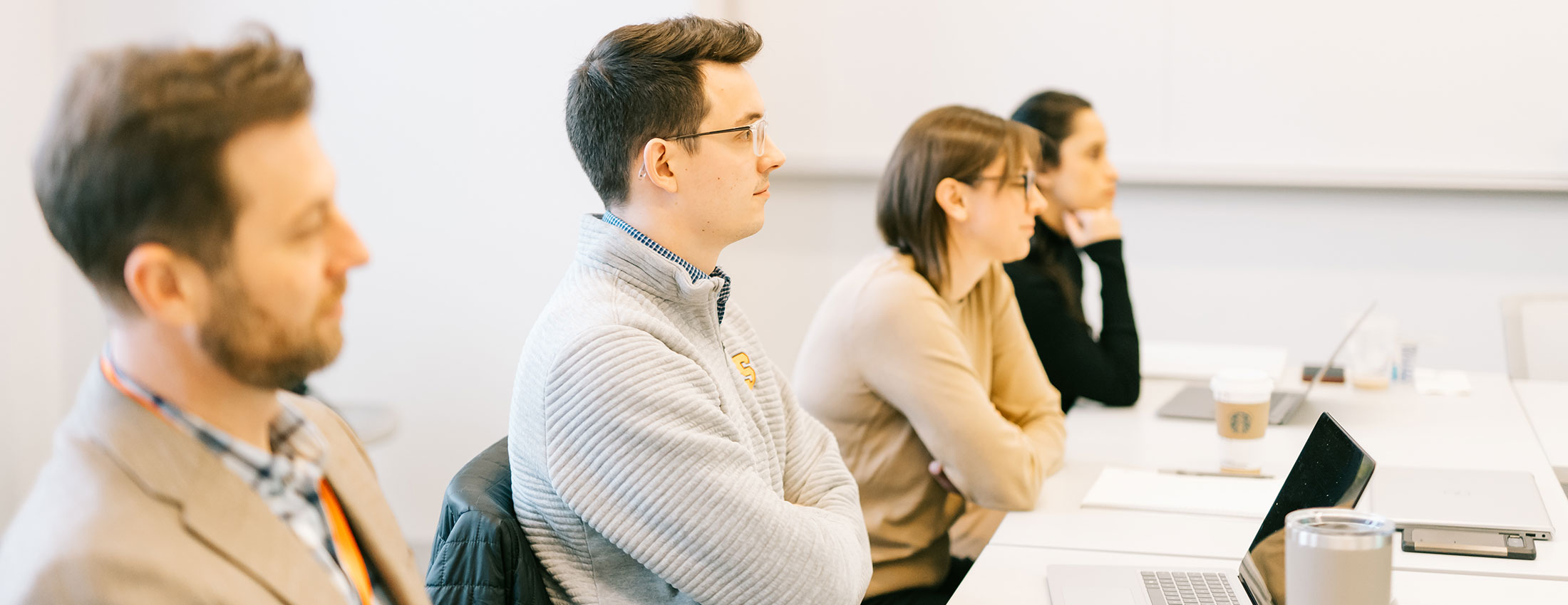 four students sit in a classroom with white walls looking forward towards their professor