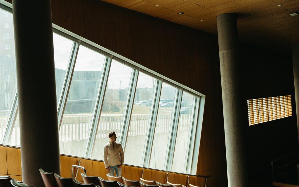 Noah looks to the side as he stands in front of large windows in a darkened courtroom