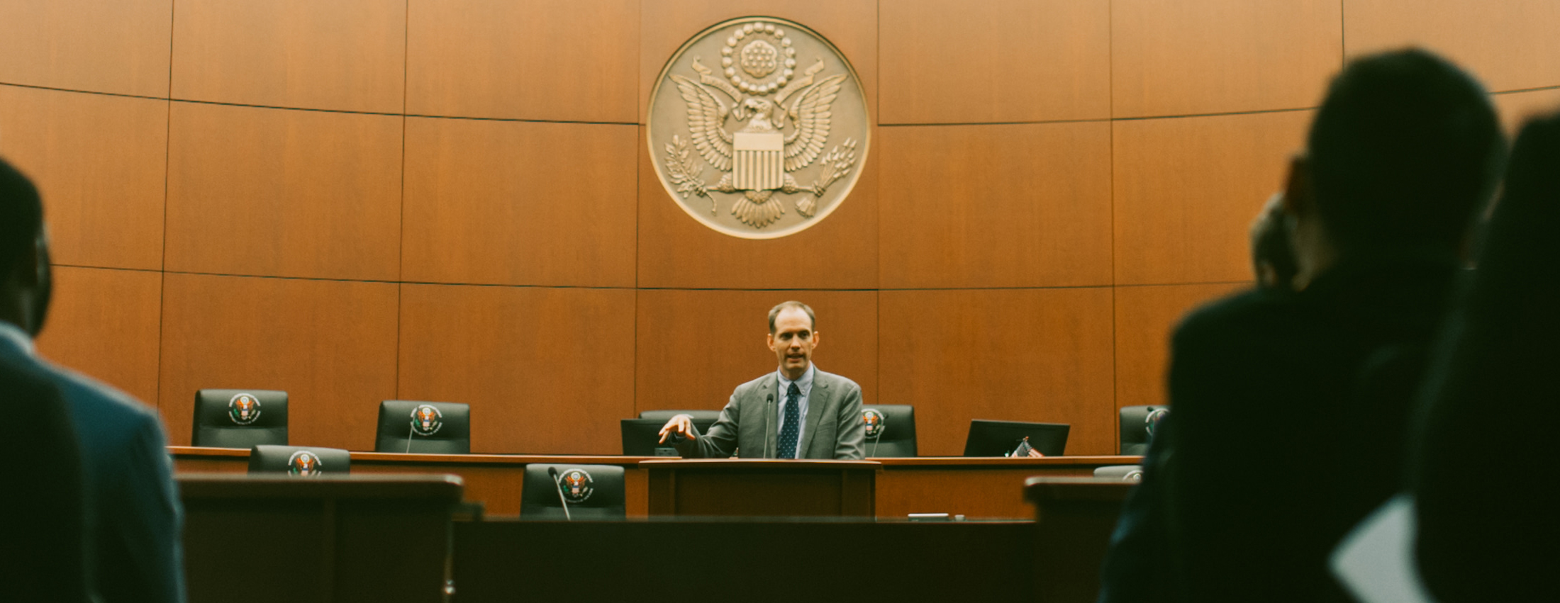 A person stands at the front of the courtroom behind a podium, onlookers are in the shadows on each side