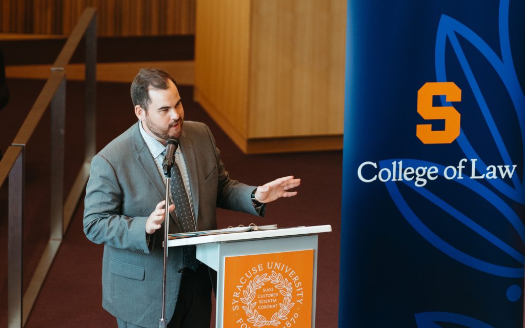 A man in a gray suit and striped tie speaks into a microphone while gesturing with his hands at a podium bearing the Syracuse University seal. A blue banner with the Syracuse University College of Law logo and an orange 