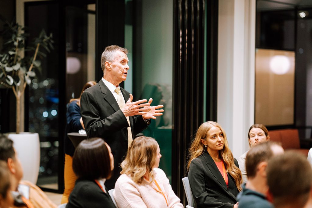 man stands behind a classroom full of students, holding up his hands and speaking to the group