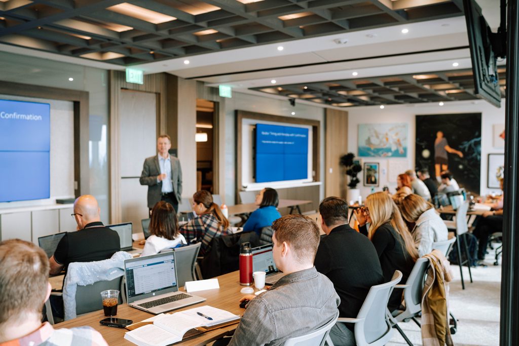 man stands in front of a classroom full of students with laptops open, teaching them