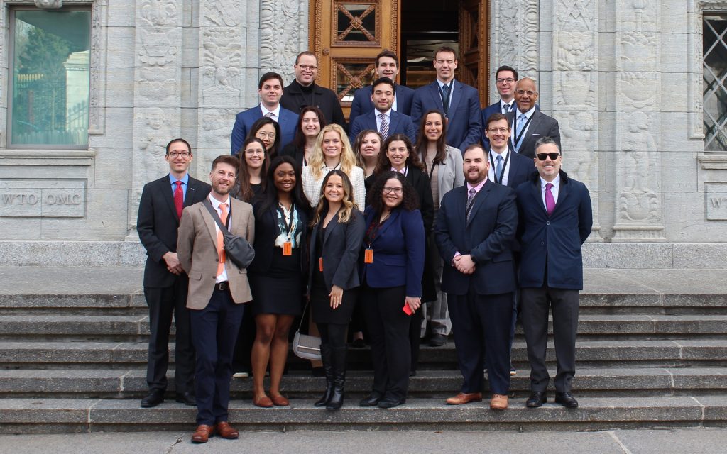 A group of professionally dressed individuals, including students and faculty, pose for a group photo on the steps of a historic stone building with intricate carvings and a wooden door. Some are wearing ID badges on orange lanyards. The engraved text 