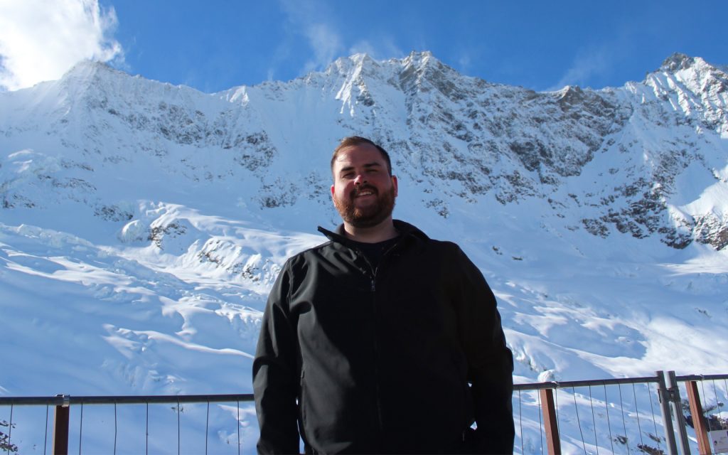 A man with a beard, wearing a black jacket, stands in front of a breathtaking snow-covered mountain range under a clear blue sky. He is smiling, and the bright sunlight highlights the rugged peaks and icy terrain. A metal railing is visible in the foreground, suggesting an observation point or scenic overlook.