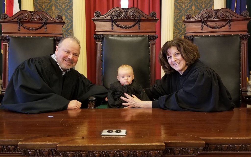 Judge Karalunas sits in a courtroom in her robe holding a baby in the middle seat next to her