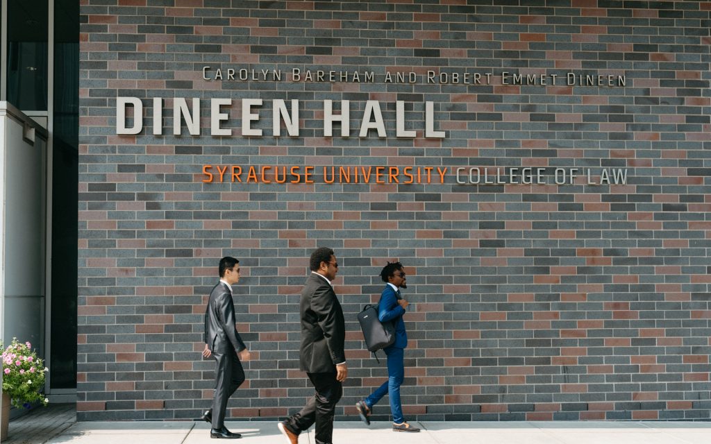 Three individuals in professional attire walking past the exterior of Dineen Hall, Syracuse University College of Law, with the building