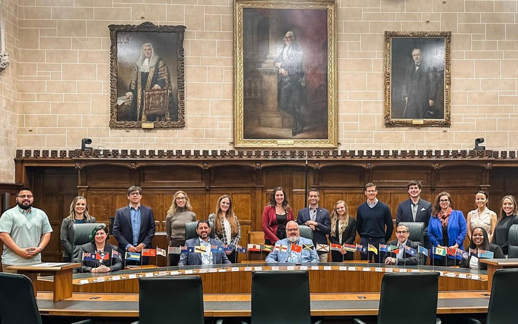 A large group of students and faculty pose for a photo at a long table. Large portraits hang on the wall in the background.