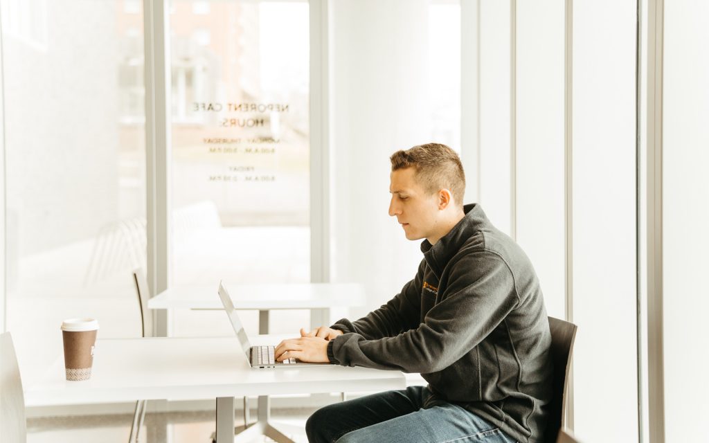 Zachary Mazuzan sits at a table in the Neporant Cafe, working on a laptop with a coffee cup in front of him on the table