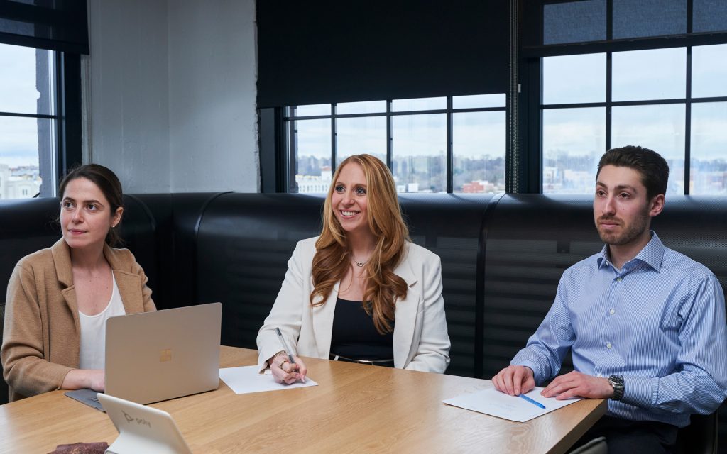 Three people sit at a conference table and listen to a speaker who is out of the frame