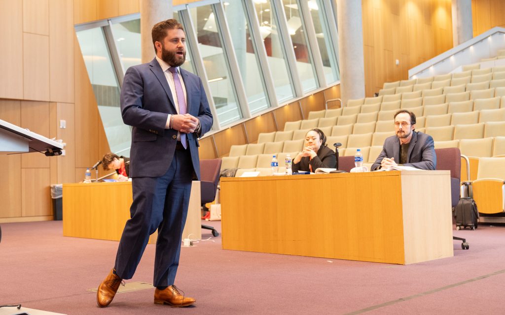 An individual in a blue suit and light purple tie stands in front of a seated audience in a lecture hall, speaking and gesturing with hands clasped. Two people sit behind a wooden desk, observing attentively. The lecture hall features modern architecture with slanted windows and beige seating.