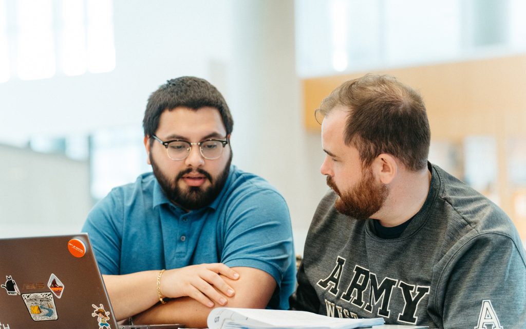 Two men sit at a table engaged in discussion, looking at each other. One wears a blue polo shirt and glasses, while the other wears a gray Army West Point sweatshirt. A laptop covered in various stickers and an open book or notebook are on the table. The background is bright and modern, suggesting a study or work environment.