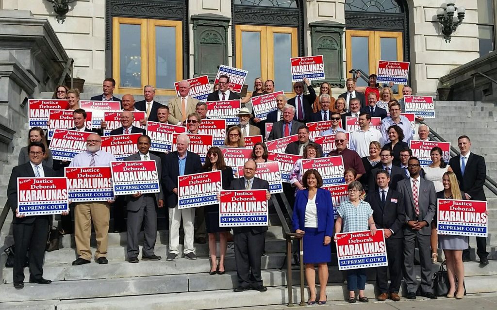 Group stands on the Courthouse steps holding up signs to re-elect Judge Karalunas, with her standing at the front smiling in a blue suit