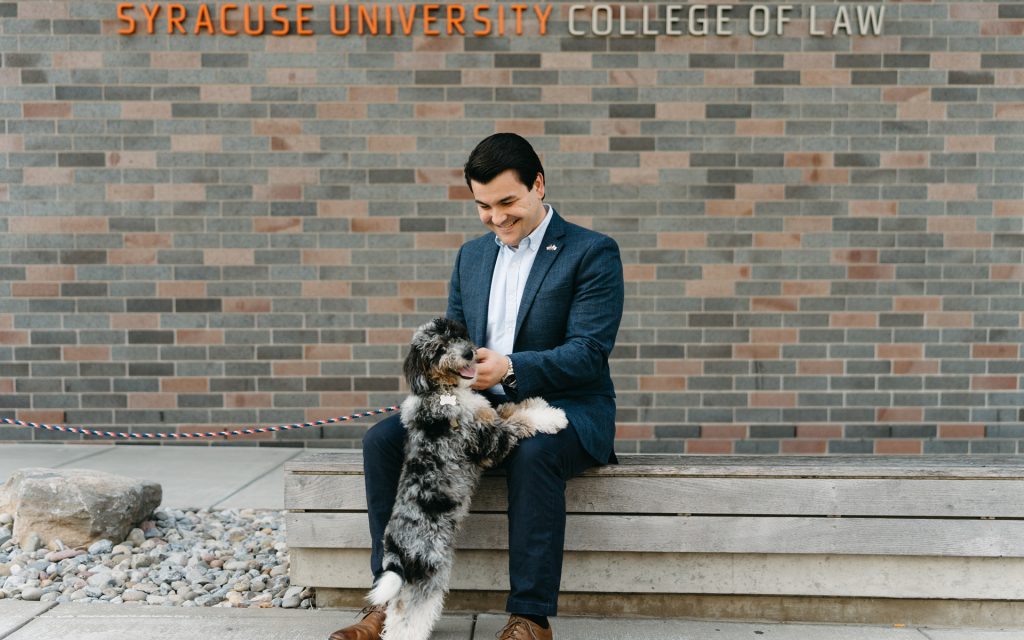 The man is sitting on a bench outside Syracuse University College of Law