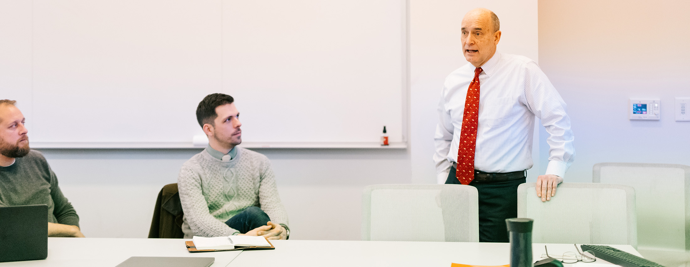 judge baker speaks to two students in a classroom, wearing an orange tie and standing in front of a white wall