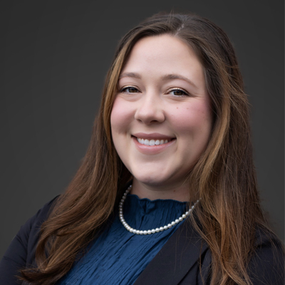 A person with long, brown hair is wearing a dark blazer over a blue top and a pearl necklace. The background is plain and dark, contrasting with the person