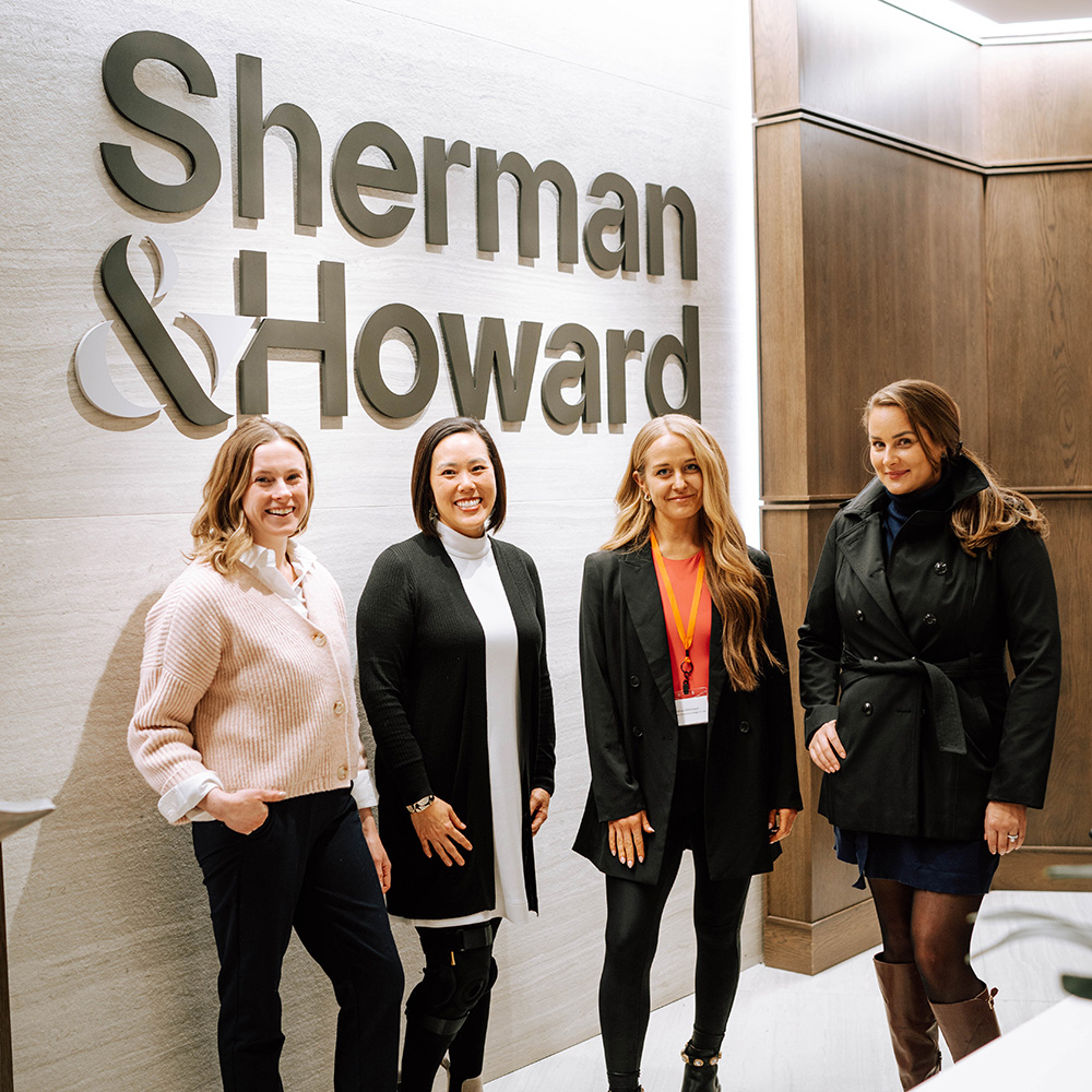 four students stand in front of a Sherman &amp; Howard sign and smile