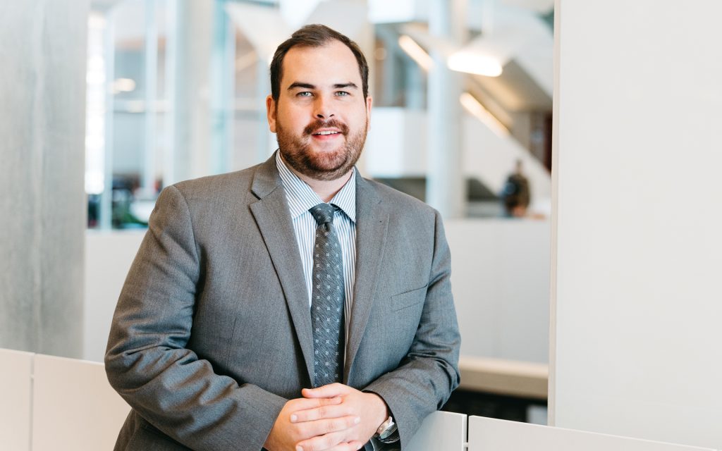 A man with short brown hair and a beard, wearing a gray suit, striped shirt, and patterned tie, smiles confidently while leaning on a white partition in a modern office or academic setting. The background features glass windows, bright lighting, and a blurred figure walking in the distance.