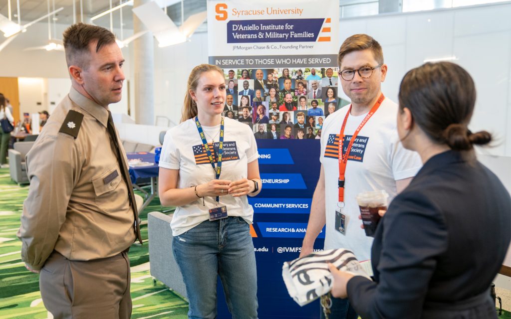 A male student in military uniform engages in conversation with two students wearing "Military and Veterans Law Society" T-shirts. Behind them is a Syracuse University D