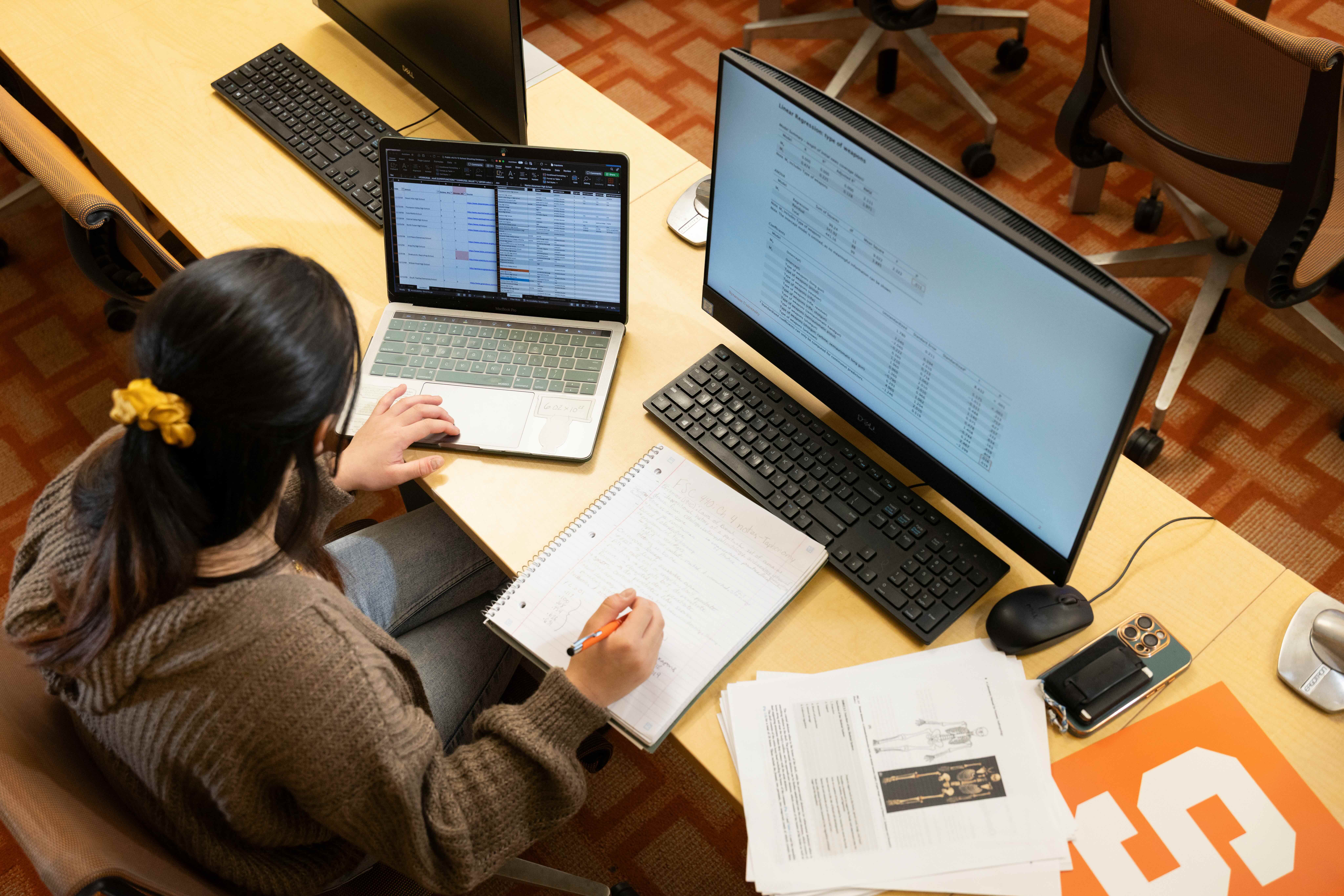 A student sits at a desk and works on their computer while also taking notes. 