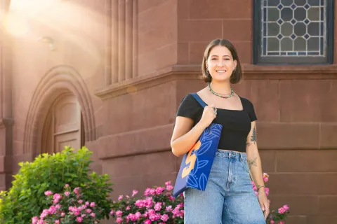 A student stands outside on campus holding a bag. 