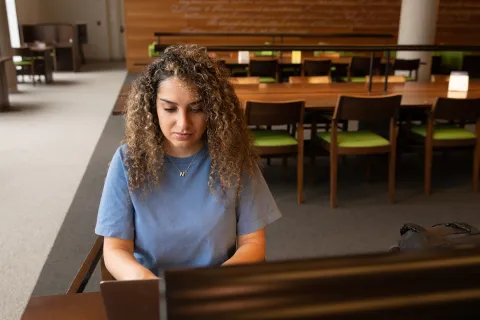 A student sitting inside a library working independently on their computer. 