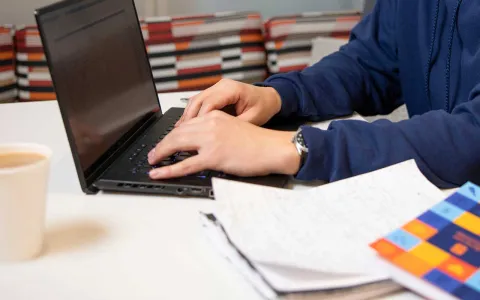 Online student typing on laptop computer with papers, notebook and cup of coffee in the foreground.