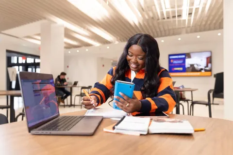 Student writing on a notebook while holding a phone with laptop open